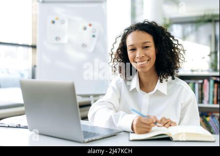 Pretty african american woman, frelancer or office worker, an intern in a company, sits at a desktop with laptop, in modern formal clothes, is trained, works, takes notes, looks at the camera, smiling Stock Photo