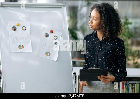 Successful focused dark-skinned woman, business analyst, marketing, stands in a modern office, near a white board, in stylish business clothes, works with charts and documents, develops a strategy Stock Photo