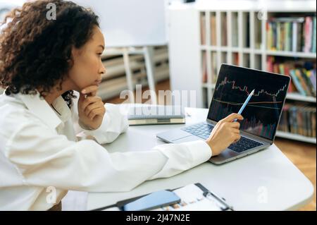 Focused African American woman, financial consultant or trader, sits in front of a computer screen, works with charts, analyzes the dynamics of cryptocurrency in the stock market, makes a calculation Stock Photo
