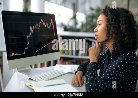 Focused smart African American woman, trader, stock broker, sits at a working table in front of computer screen, concentrated looks at the quotes charts, analyzes the dynamics and further dynamics Stock Photo