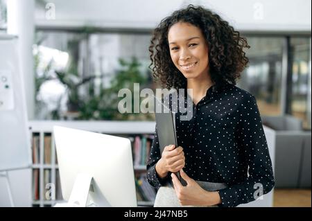 Charming young adult African American woman, manager, financial consultant, auditor, stands in modern office, in stylish business clothes, holding working papers in her hands, smiles friendly Stock Photo