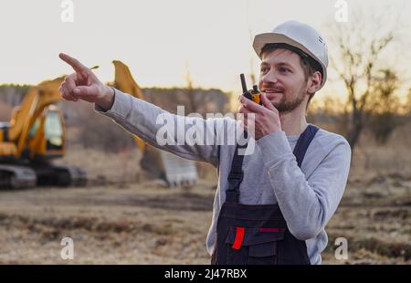 Worker in helmet talking by radio transmitter on construction site. Excavator on the background. Stock Photo