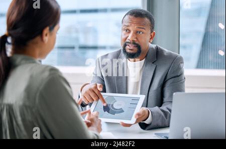 Pointing out problems based on market research. Shot of a mature businessman speaking to a colleague while analysing graphs on a digital tablet in an Stock Photo