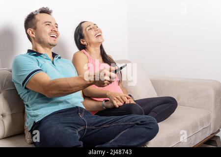 latin man and woman couple watching tv, laughing and using remote control Stock Photo