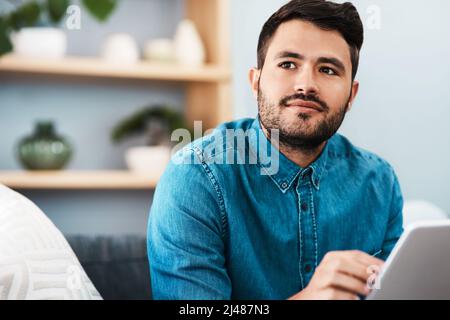 Whats the name of that app again. Cropped shot of a handsome young man looking thoughtful while using a digital tablet in his living room at home. Stock Photo