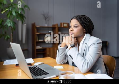 Young thoughtful african female employee looking out window at workplace and dreaming about vacation Stock Photo