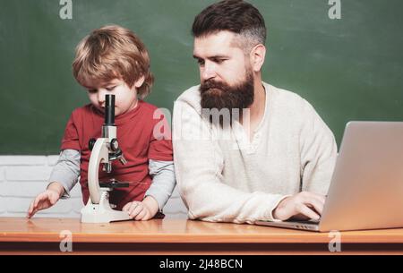 Teacher helping pupils studying on desks in classroom. Young boy doing his school homework with his father. Chalkboard background. Dad son are Stock Photo