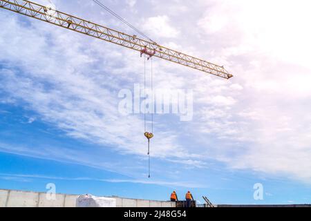 A large yellow tower crane is working against a blue sky, slingers in helmets and orange vests are working at a height, selective focus Stock Photo