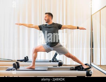 Fit man performing a lunge and stretch Warrior 2 yoga pose on a pilates reformer bed to stretch and strengthen his hip and chest muscles and promote s Stock Photo