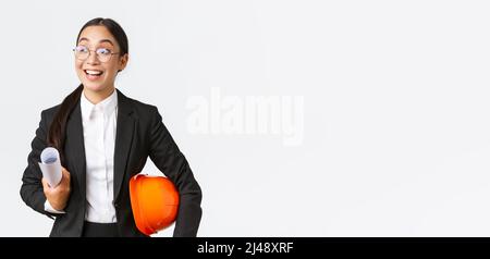 Hopeful and excited asian female architect looking around with fascination and amazement, holding blueprints and helmet, walking in construction area Stock Photo