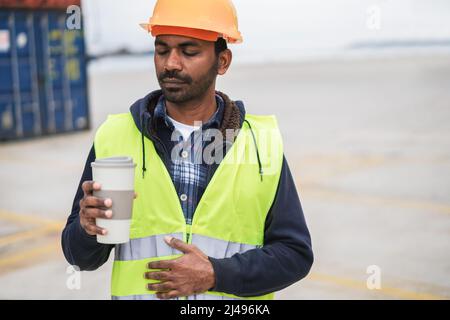 Indian worker drinking coffee with shipping containers on background at industrial port - Focus on face Stock Photo