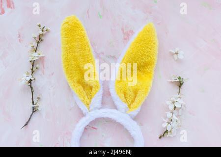 Yellow and white bunny ears and tree brunches with white cherry blossom flowers on a pink background. Easter celebration concept. Top view, flat lay, copy space Stock Photo