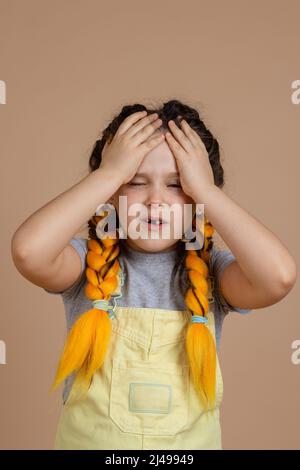 Small female child with yellow kanekalon braids, having headache touching head with hands and with half-opened mouth and eyes in yellow jumpsuit and Stock Photo