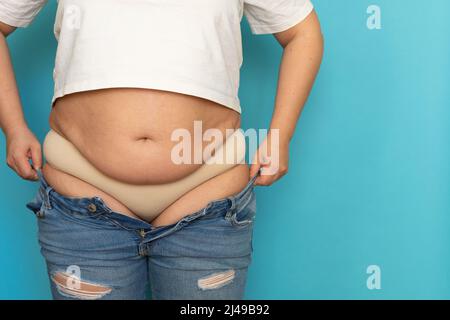 Cropped photo of plump woman wearing white short T-shirt, standing on blue background and trying to pull on blue jeans. Stock Photo