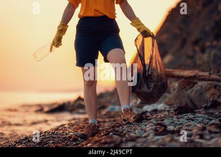 Cleanup garbage on the ocean coast. A volunteer holds a plastic bottle and poses on the beach. Close-up of the legs. Sunset in the background. The con Stock Photo