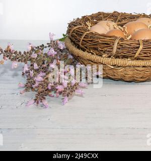 Farmers' Eggs. Copy Space. White background. Brown fresh  eggs in wicker nest with straw on a flat table. Wildflowers on the side. Stock Image. Stock Photo