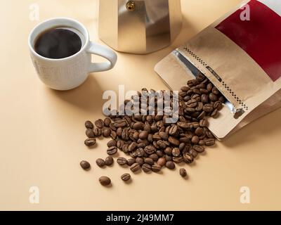 Cup of freshly brewed coffee and roasted beans on a beige background. Making tasty strong coffee. Whole organic coffee beans spilled out of paper bag. Stock Photo