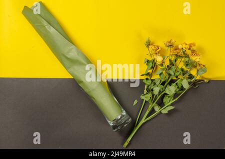 Fading flowers against a colored background. A branch of small yellow roses. Roll of green paper inside the transparent broken glass. Gray and yellow Stock Photo