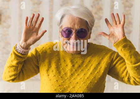 Funny caucasian grandmother in purple sunglasses looking at camera and smiling, and holding her hands up. Vintage wallpaper in the background. High quality photo Stock Photo