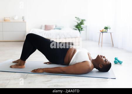 Curvy black lady stretching her legs, exercising on yoga mat