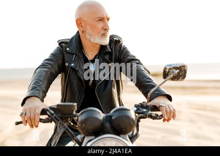 Bold senior man wearing leather jacket riding motorcycle outdoors on summer day Stock Photo