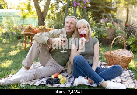 Excited elderly spouses celebrating anniversary, drinking wine, sitting on blanket while having picnic in their garden Stock Photo