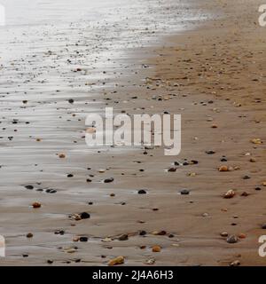 Pebbles of various types, shapes and sizes in the sand, with light reflecting on the water as it meets the shore. Sandsend beach, Yorkshire Stock Photo