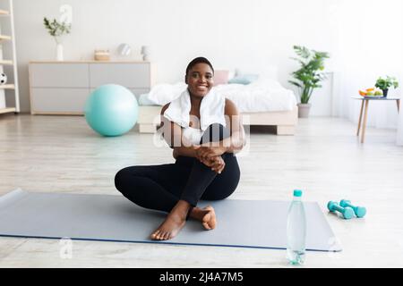 Smiling overweight young black woman sitting on yoga mat, resting after home fitness, copy space Stock Photo