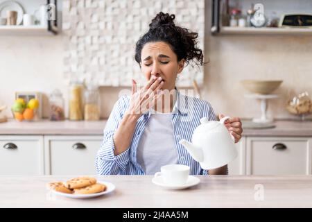 Sleepy tired woman waking up early, pouring tea into cup and yawning, sitting in modern kitchen interior, copy space Stock Photo