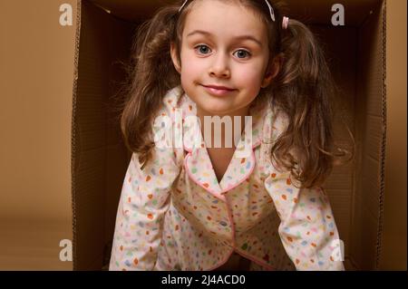 Close-up baby girl with two ponytails in pajamas peeking out from behind a cardboard box, isolated over beige background with copy space. Home, housin Stock Photo