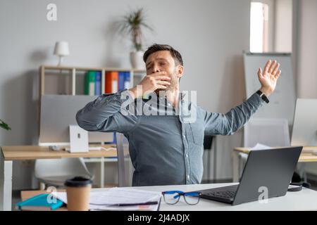 Sleepy mature businessman yawning, tired of working with laptop in office, male workaholic feeling sleepy or bored Stock Photo