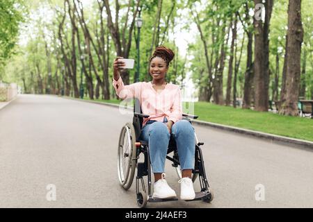 Full length of happy young black woman in wheelchair taking selfie on cellphone at park, copy space Stock Photo