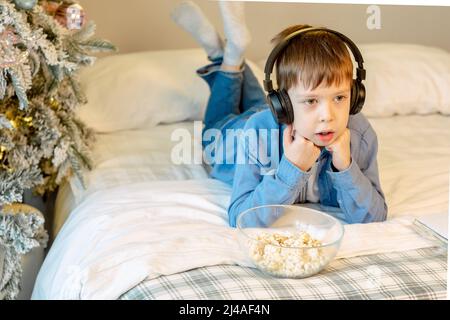 Cute boy with bluetooth headphones eating popcorn while lying in