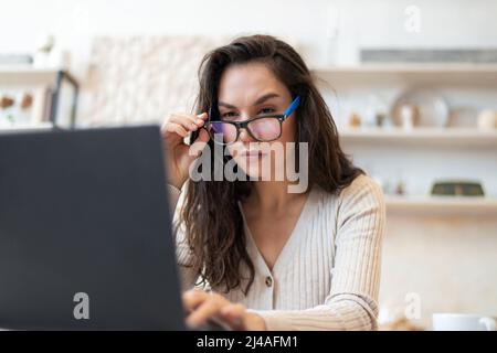Stressed freelancer woman looking at laptop screen while working remotely from home, sitting in kitchen with computer Stock Photo