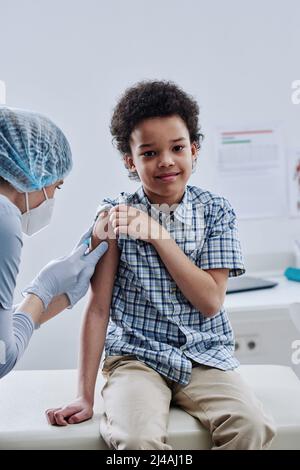 Portrait of African little boy smiling at camera while nurse giving injection in his arm at procedure room Stock Photo