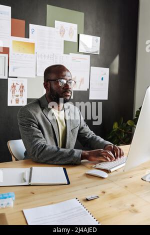 African male doctor in suit sitting at his workplace in front of computer monitor and working online at office Stock Photo