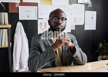 Portrait of African male doctor in eyeglasses looking at camera while sitting at table and working at office Stock Photo