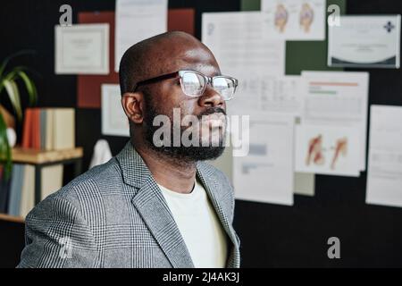 African medical specialist in eyeglasses thinking over the work while looking at window at office Stock Photo