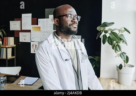 African male therapist in white coat and eyeglasses looking at window with smile on his face during work at office Stock Photo