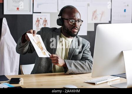 African traumatologist in headphones pointing at picture with body parts and teaching online using computer at office Stock Photo