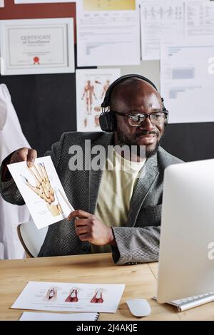 African male doctor in headphones pointing at picture with human hand and talking to his colleagues during online meeting on computer Stock Photo