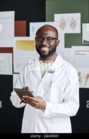 Portrait of African successful doctor in white coat smiling at camera while using tablet pc during work at office Stock Photo