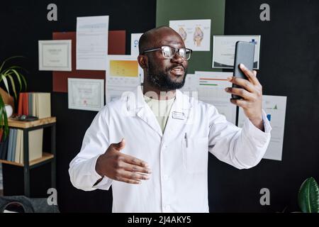 African male doctor in white coat using mobile phone for online consultation during work at office Stock Photo