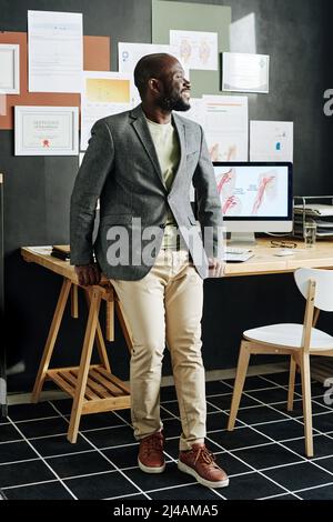 Successful African physician smiling and looking away while standing at his office with computer and certificates Stock Photo