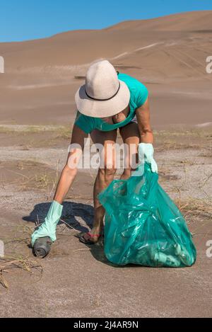 Unrecognizable mature woman in white cap and green gloves bending over with a bag of garbage. Picking up an abandoned soda can from the sand. Stock Photo