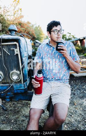 Young latin man drinking mate and relaxing, in a rural space, beside an old little blue tractor. Latin beverage. Stock Photo