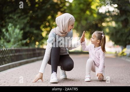 Front view of happy likable Muslim family, mother and daughter, squatting down on the park track, ready to run, having fun and giving high five each other Stock Photo
