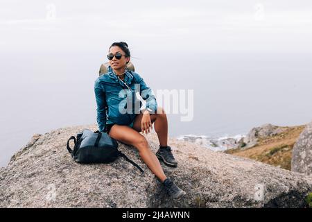 Fit woman in sportswear sitting on a rock during hike. Young female in sunglasses enjoying the view from the peak. Stock Photo