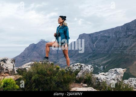 Smiling woman hiker with camera and backpack standing on the top of the mountain. Happy female stepping up on the rock. Stock Photo