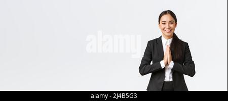 Hopeful glad asian businesswoman in black suit bowing politely and say namaste, greeting clients and smiling friendly. Thankful female manager Stock Photo